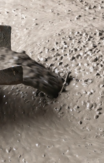Das Bild zeigt frischen, sehr flüssigen Beton, der aus einer Rinne auf eine Baustelle gegossen wird. Die Betonmasse ist glänzend und weist eine hohe Fließfähigkeit auf, einzelne grobe Gesteinskörner sind deutlich sichtbar. Die Oberfläche wirkt nass und glatt, was typisch für leicht verdichtbaren Beton ist, der sich ohne große Rüttelarbeit gleichmäßig verteilt.

                                    
